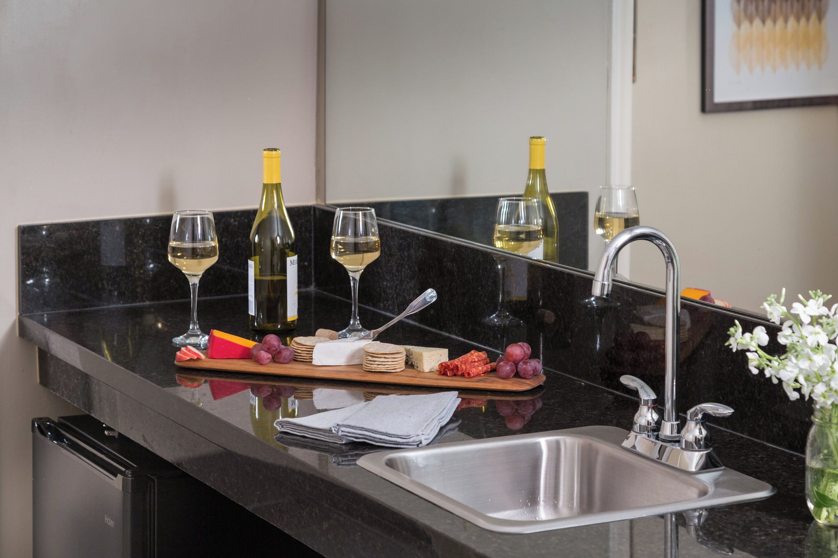 Una encimera de cocina con un lavabo, copas de vino, botellas, queso y uvas.
