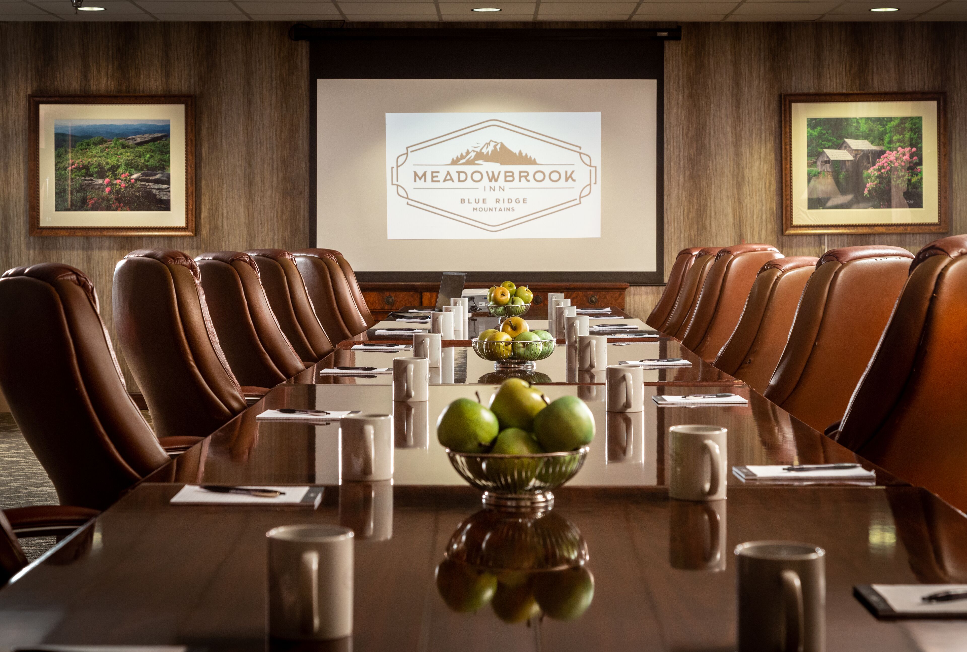 A conference room with a long table, brown leather chairs, a bowl of fruit, and a projector screen displaying the Meadowbrook Inn logo.