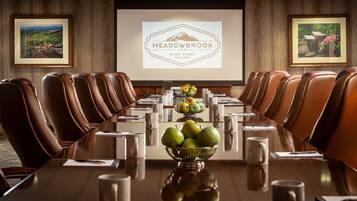 A conference room with a long table, brown leather chairs, a bowl of fruit, and a projector screen displaying the Meadowbrook Inn logo.