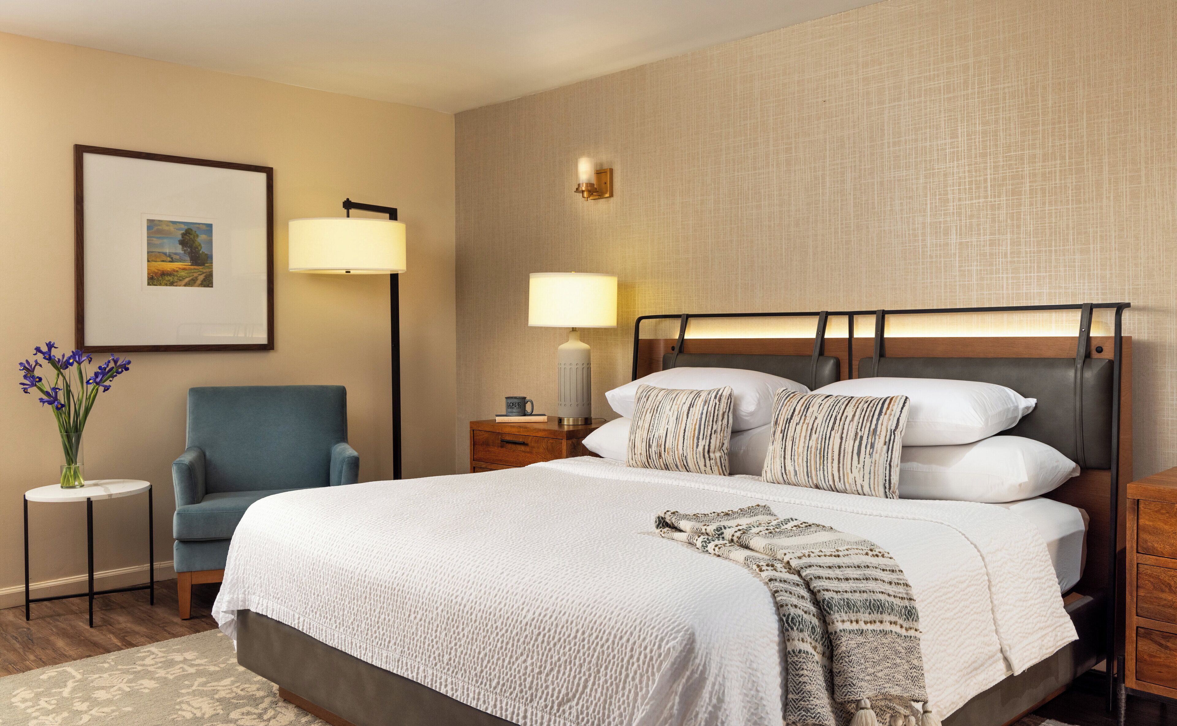 A neatly arranged hotel room with a bed, a chair, a nightstand, a lamp, and a framed picture on the wall.