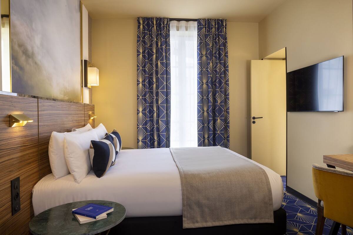 A hotel room with a bed, a bedside table with books, a TV, and a window with patterned curtains.