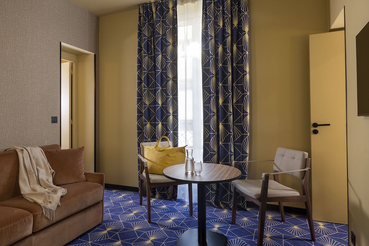 A hotel room with a brown sofa, a round table with chairs, and a patterned carpet.