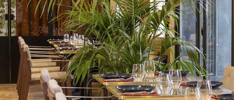A dining area with tables set for service, featuring a central plant and a view of the city through large windows.