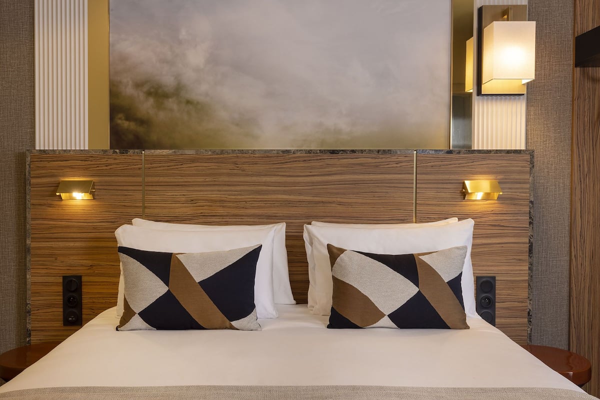Habitación de hotel con cabecera de madera, dos almohadas decorativas, un cuadro abstracto grande e iluminación en la pared.