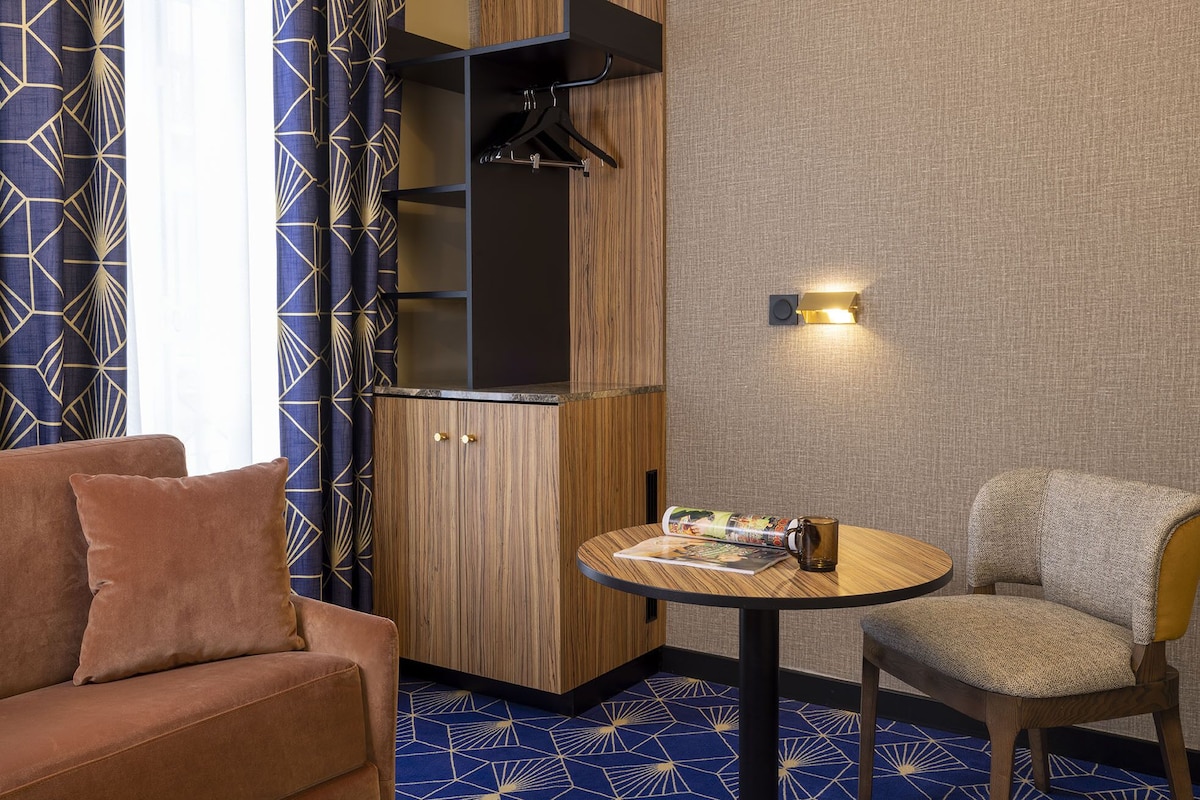 A compact room with a sofa, a round table with a magazine and a mug, a wooden cabinet with a hanging rack, and a wall-mounted light fixture.