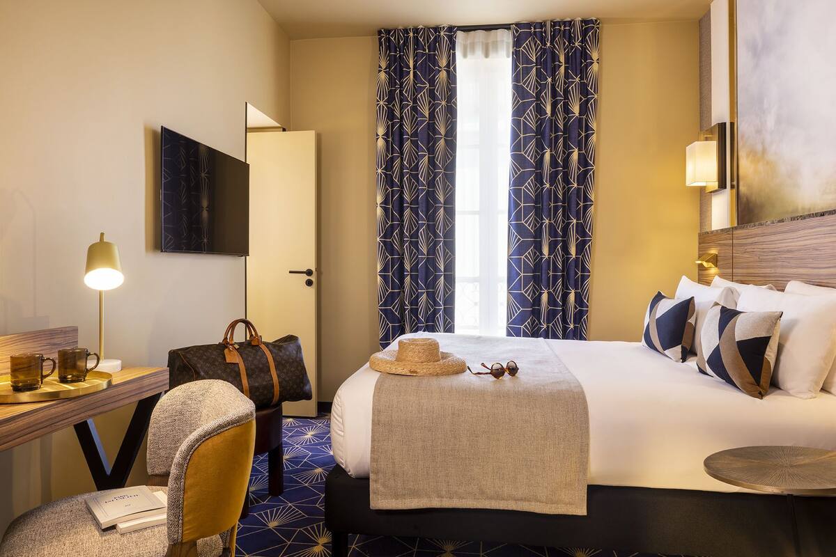 Una habitación de hotel con una cama, un escritorio con lámpara, una silla y una maleta.