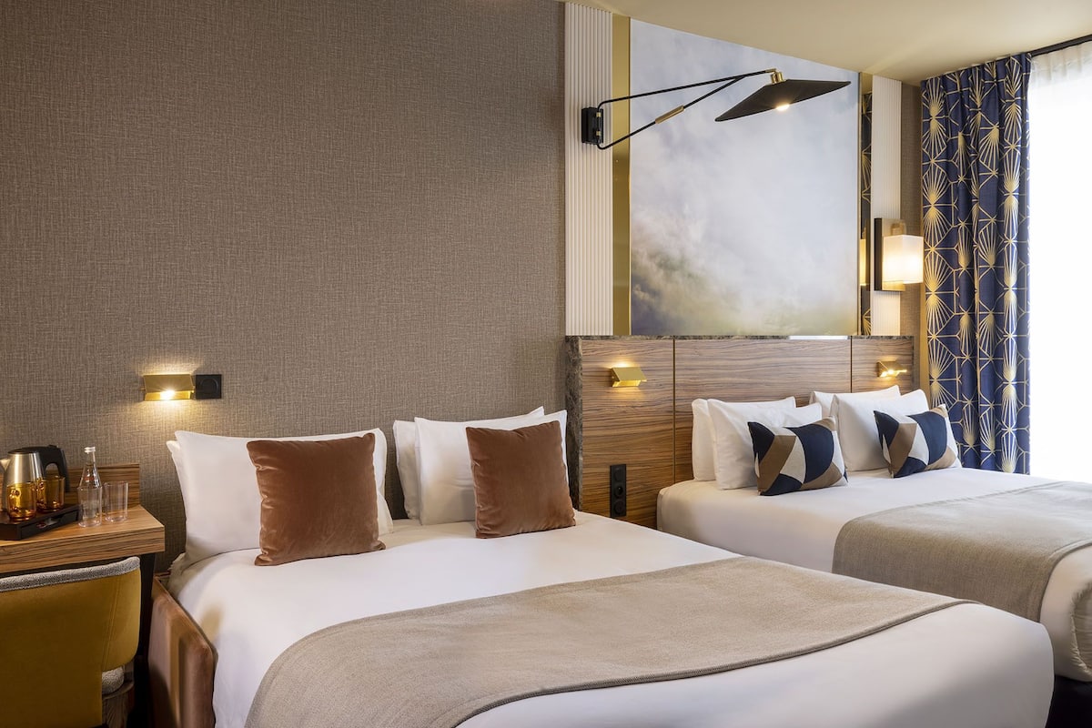 Habitación de hotel con dos camas, cabecera de madera, mesita de noche con lámpara y un ventanal con cortinas.