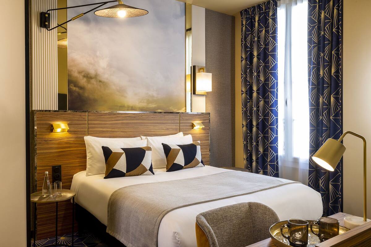 A hotel room with a large bed, bedside lamps, a small table with glasses, and a decorative wall piece.