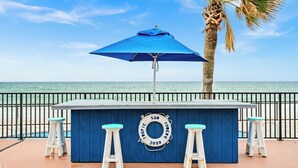 A blue bar with white stools and a blue umbrella, located by a beach with a palm tree and a railing. - Tropic Sun Towers (Ormond Beach)