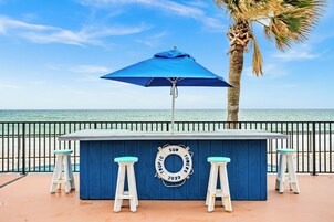 Un bar bleu avec des tabourets blancs et un parasol bleu, situé près d’une plage, avec un palmier et une rambarde.