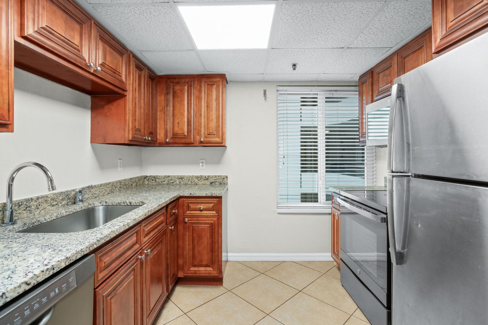A kitchen with granite countertops, wooden cabinets, a stainless steel refrigerator, and a window with blinds.