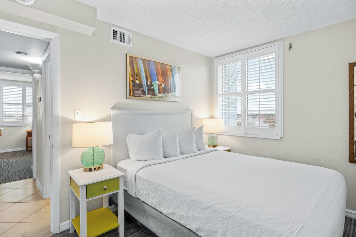 A hotel room with a large bed, a bedside table with a lamp, a painting on the wall, and a window with blinds.