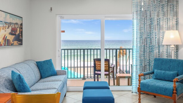A room with a balcony overlooking the beach, a sofa, a chair, and a table.