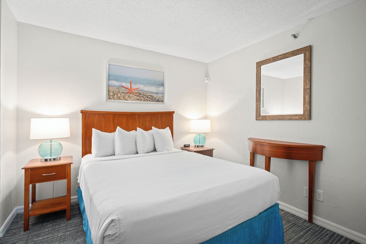 A hotel room with a large bed, two bedside tables with lamps, a wooden headboard, a wall-mounted mirror, and a framed picture above the bed.