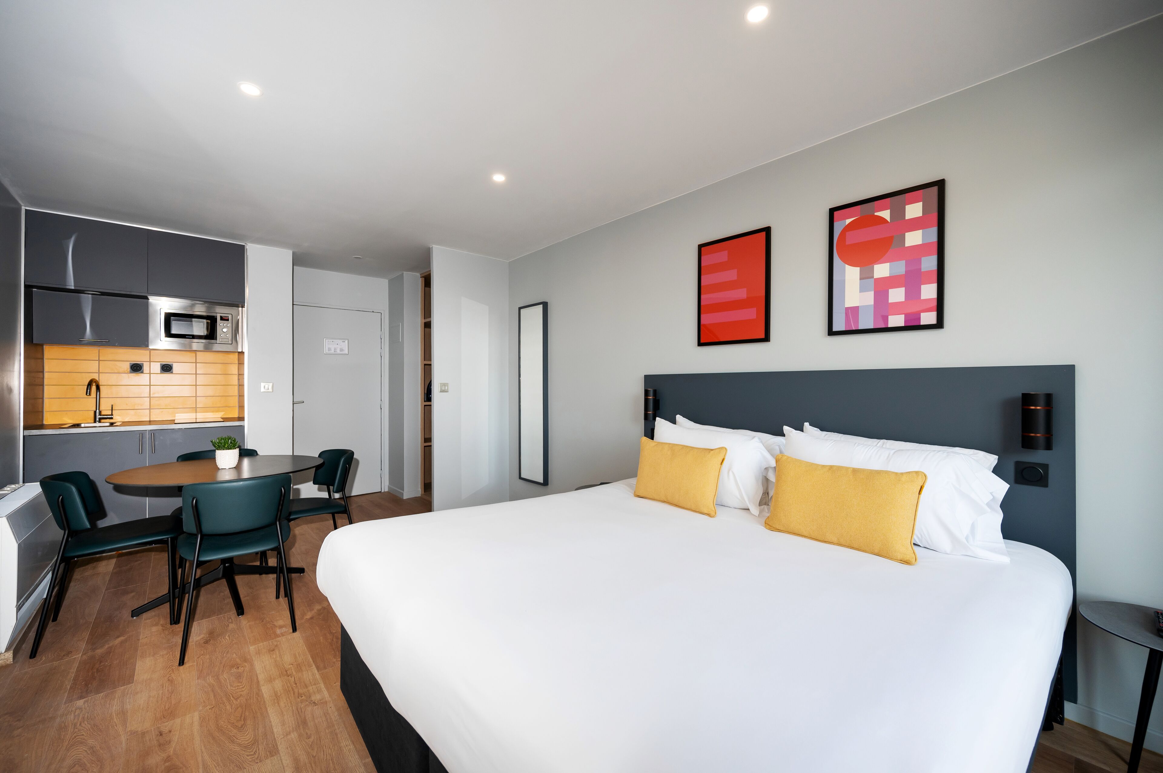 A hotel room with a bed, a dining table with chairs, a kitchenette, and a bathroom.