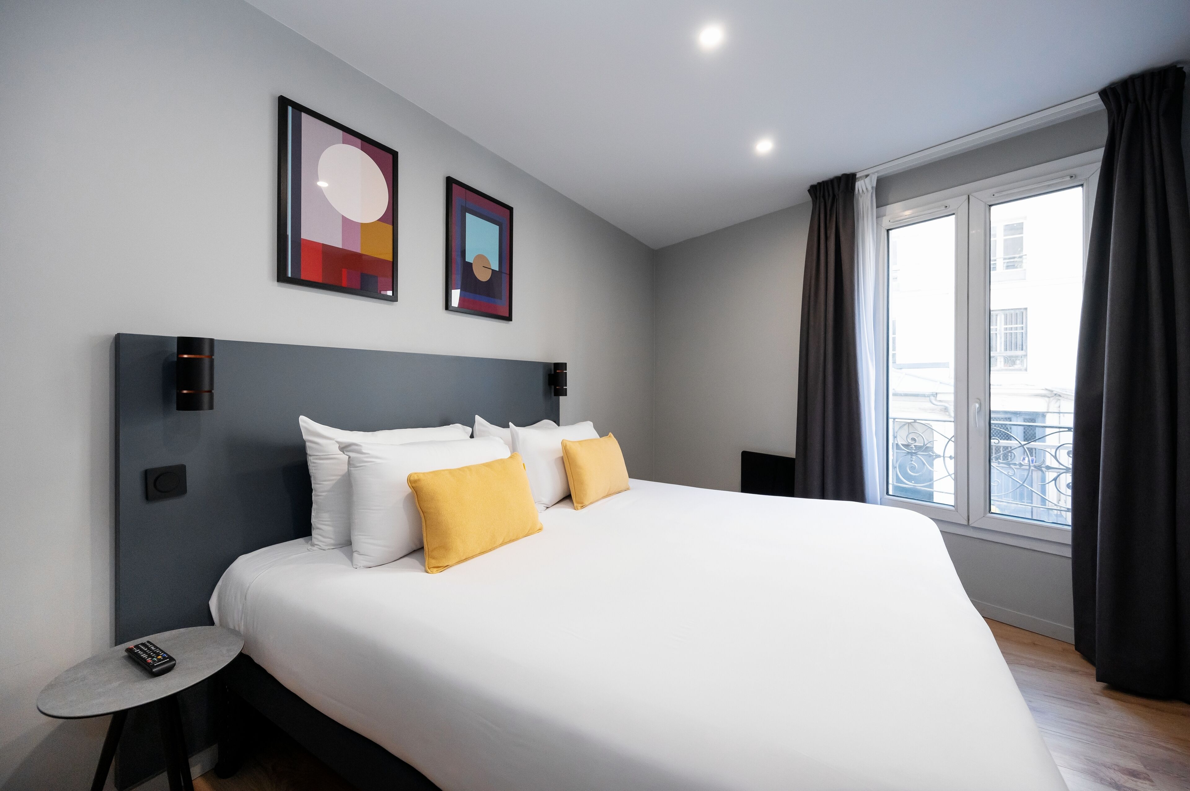 A modern hotel room with a large bed, two framed artworks on the wall, a small table with a remote control, and a window with curtains.