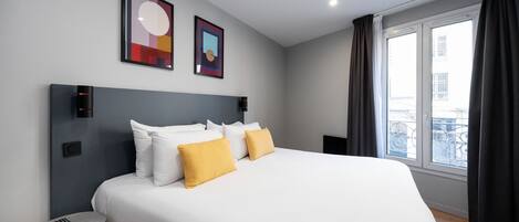 A modern hotel room with a large bed, two framed artworks on the wall, a small table with a remote control, and a window with curtains.