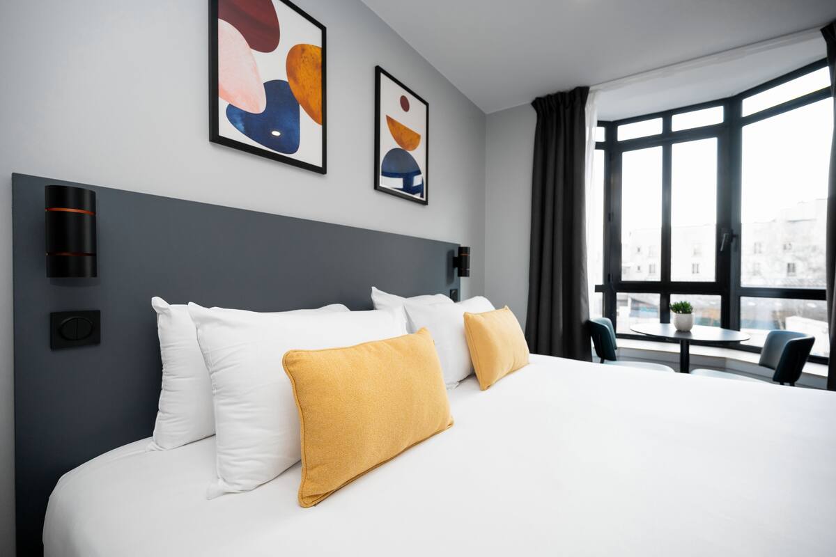 Een moderne hotelkamer met een groot bed, witte en mosterdkleurige kussens, abstracte kunst aan de muren, een raam met uitzicht op gebouwen en een klein tafeltje met stoelen.