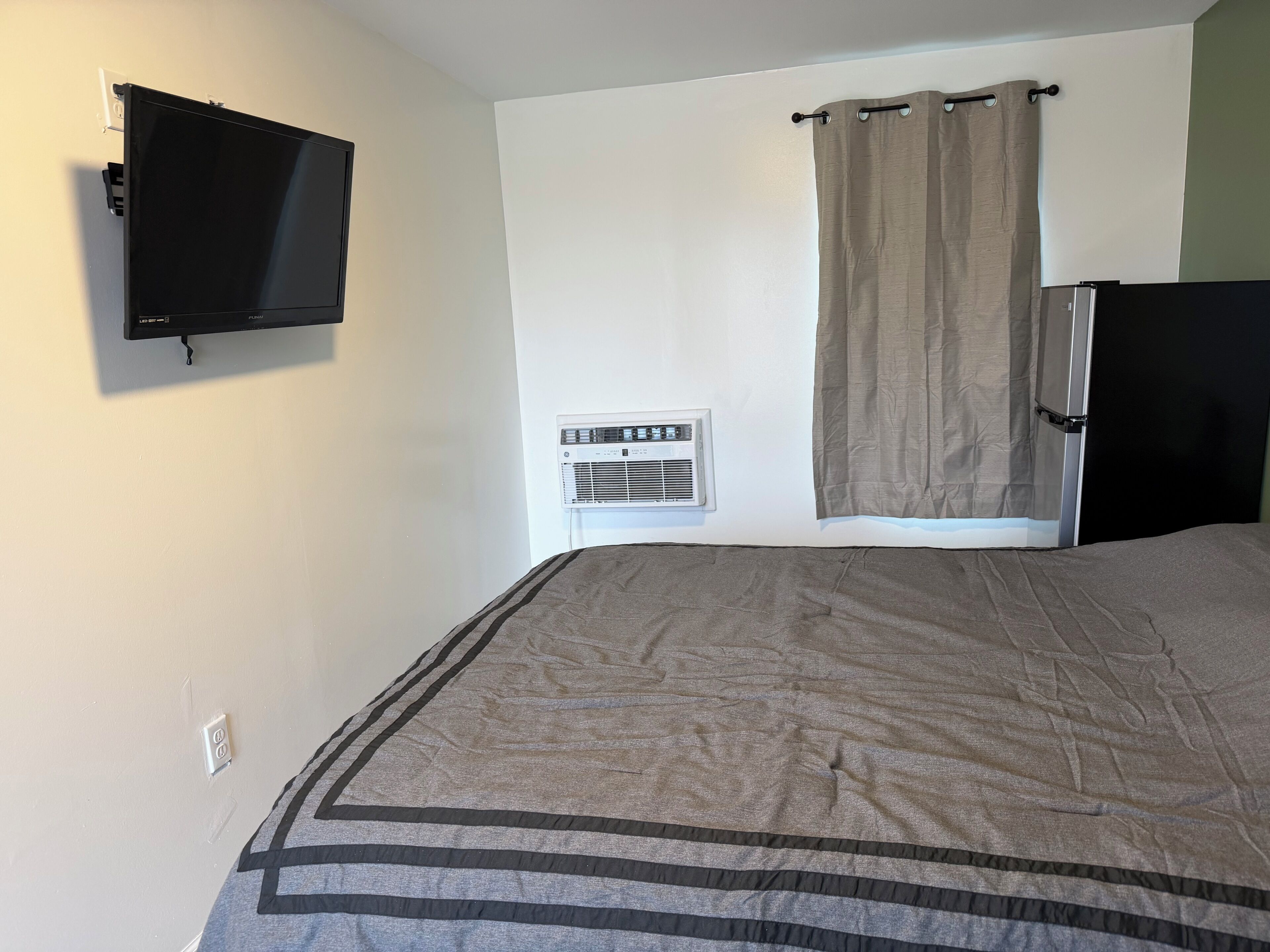 A bedroom with a bed, a wall-mounted TV, an air conditioning unit, and a window with a curtain.