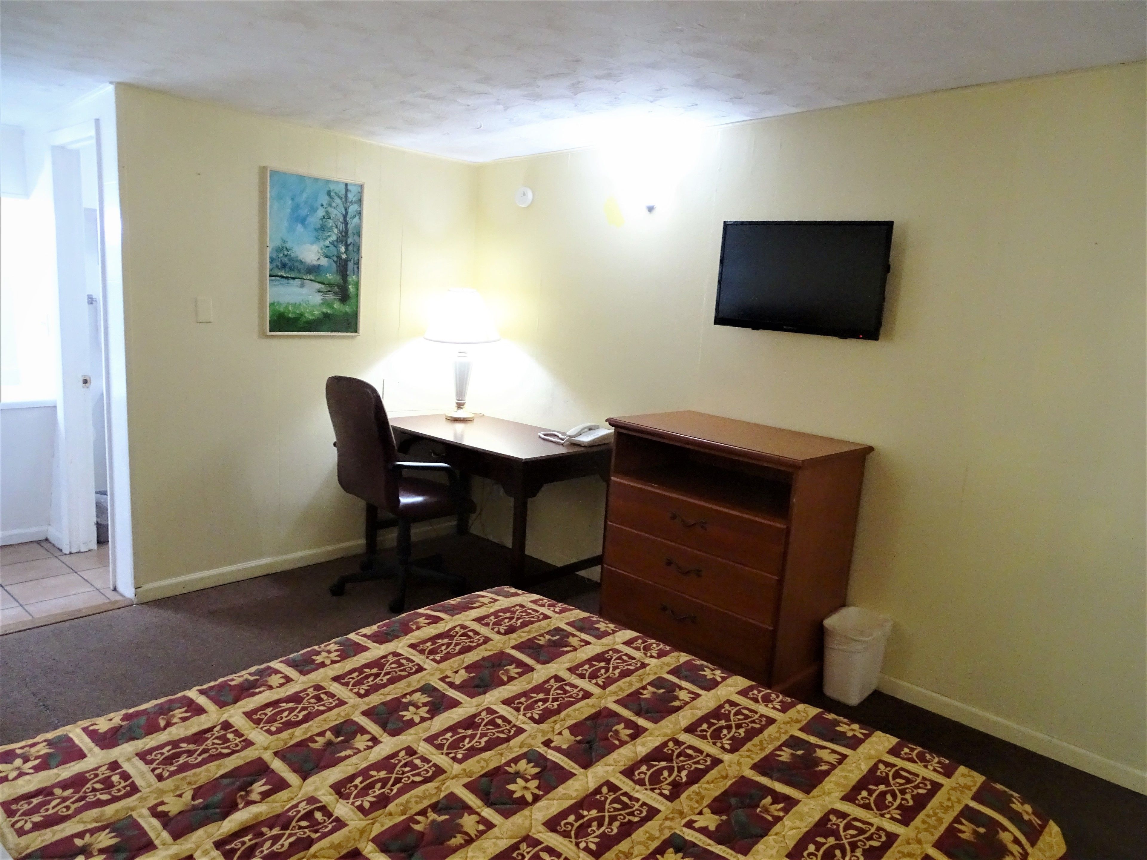 A bedroom with a bed, a desk, a chair, a dresser, a TV, and a painting on the wall.