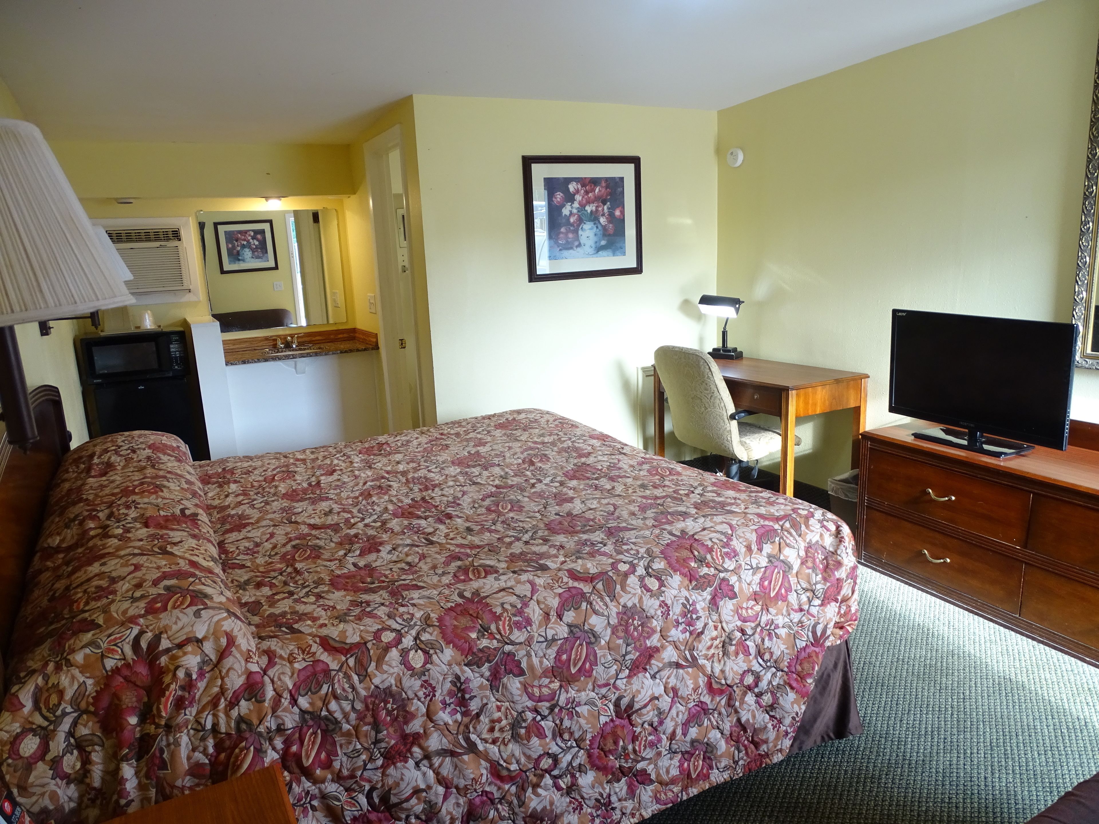 Une chambre d’hôtel avec une couvre-lit à motifs floraux, un bureau avec une télévision, une chaise, une lampe et un four à micro-ondes.