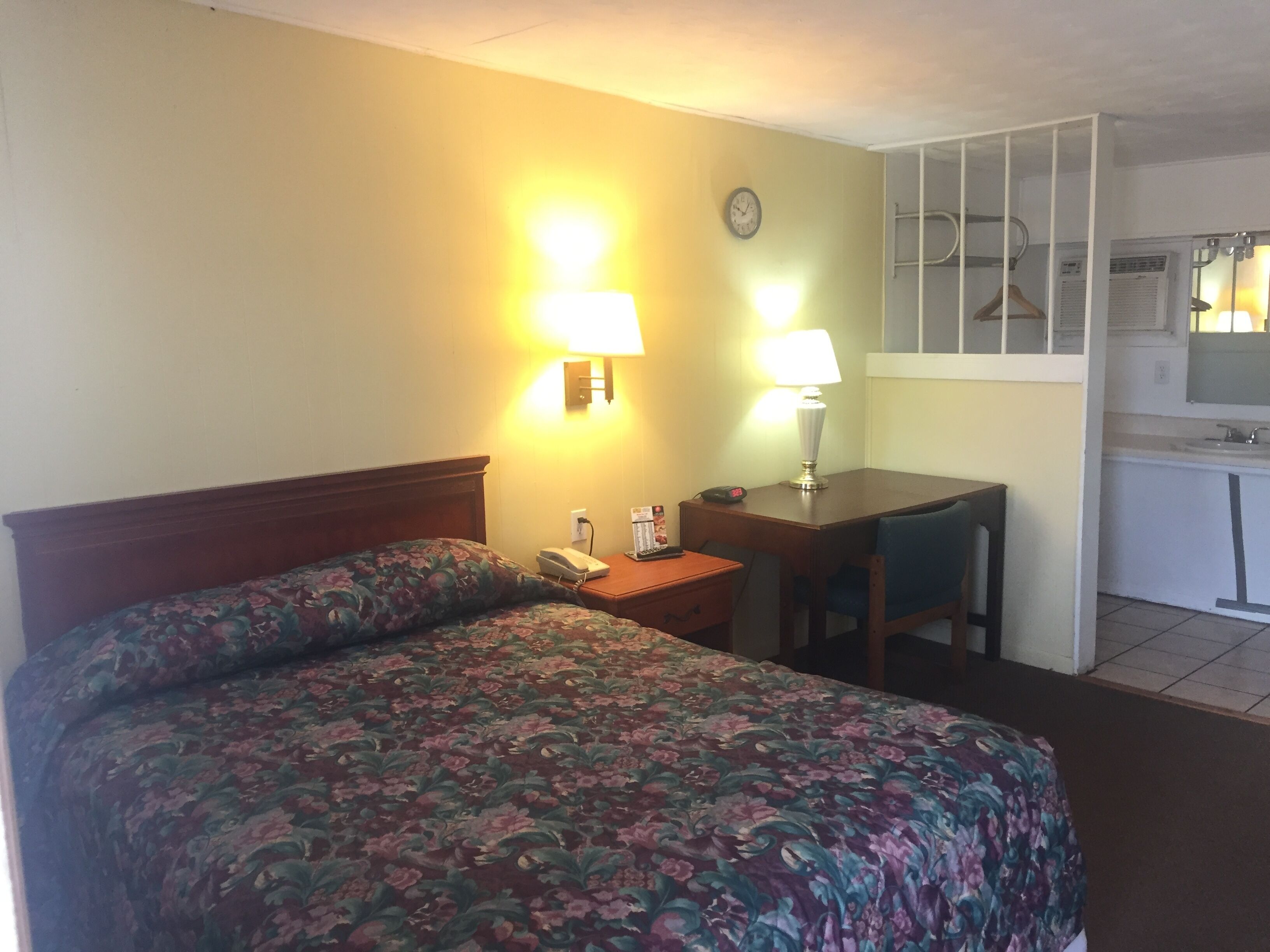 Ein Hotelzimmer mit Bett, Schreibtisch, Stuhl, Lampe, einer Uhr und einer Küchenzeile.