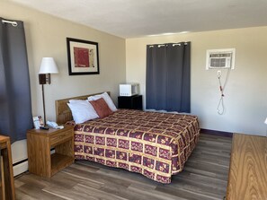 A hotel room with a wooden bed, a nightstand with a lamp, a TV, and a wall-mounted air conditioner.
