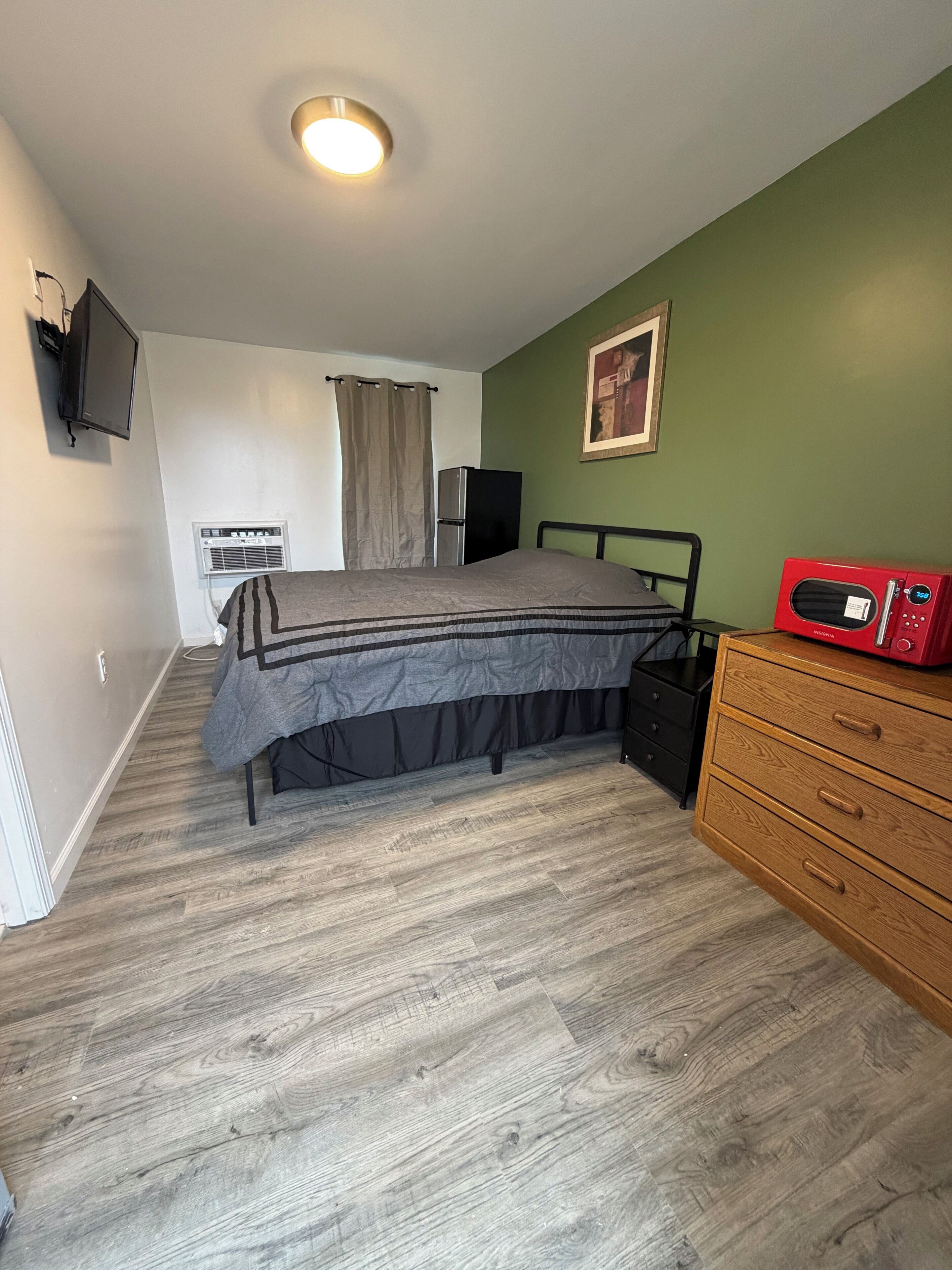 A bedroom with a bed, a dresser, a TV, an air conditioner, and a microwave.