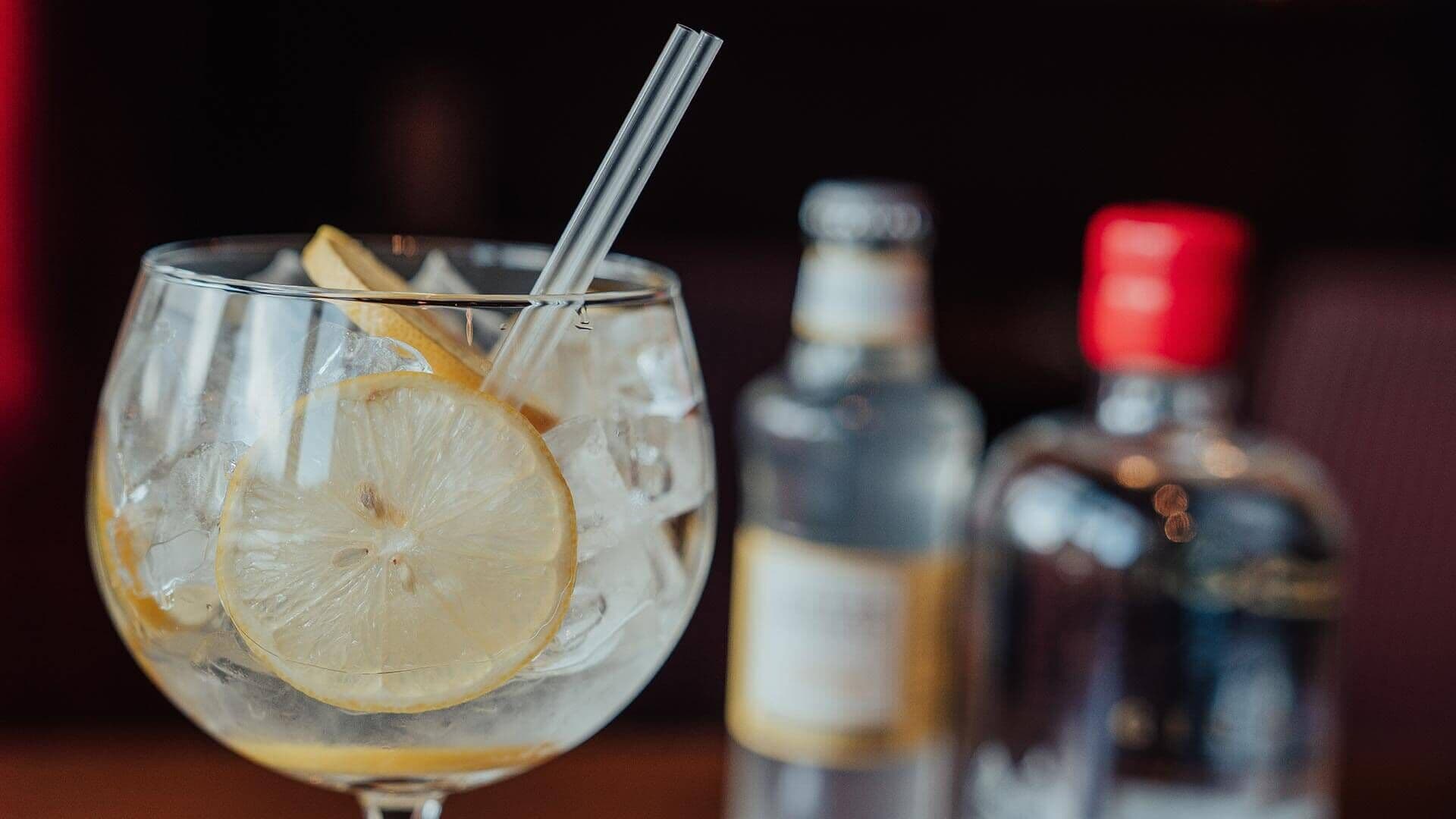 A glass with ice and a lemon slice, a straw, and two blurred bottles in the background.