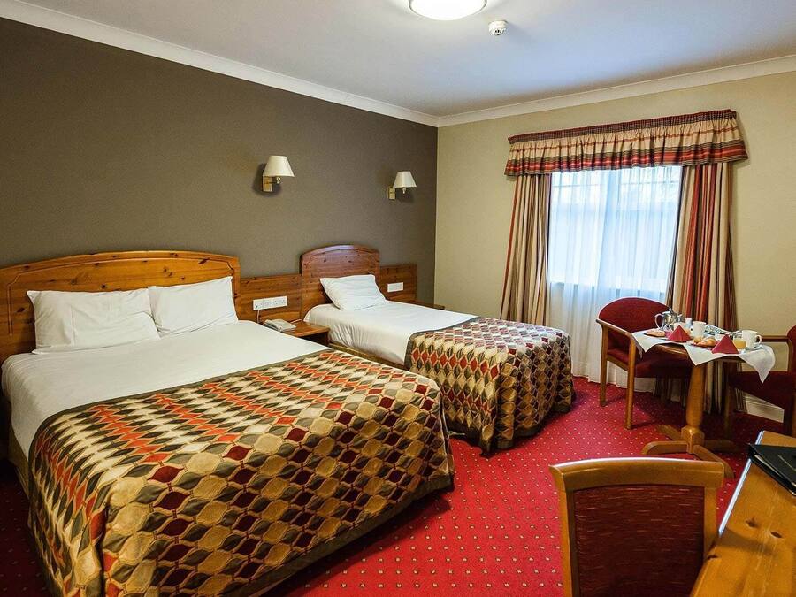 A hotel room with two single beds, a small table with a chair, and a window with curtains.