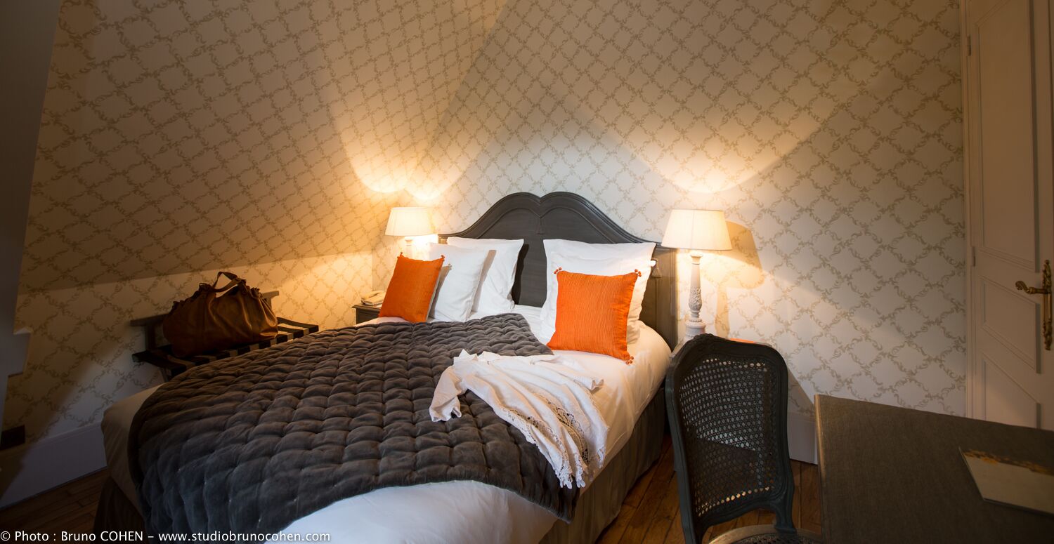 A neatly made bed with a dark quilt, two pillows, and a blanket. There are two bedside lamps, a brown handbag, and a chair.