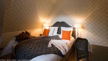 A neatly made bed with a dark quilt, two pillows, and a blanket. There are two bedside lamps, a brown handbag, and a chair.