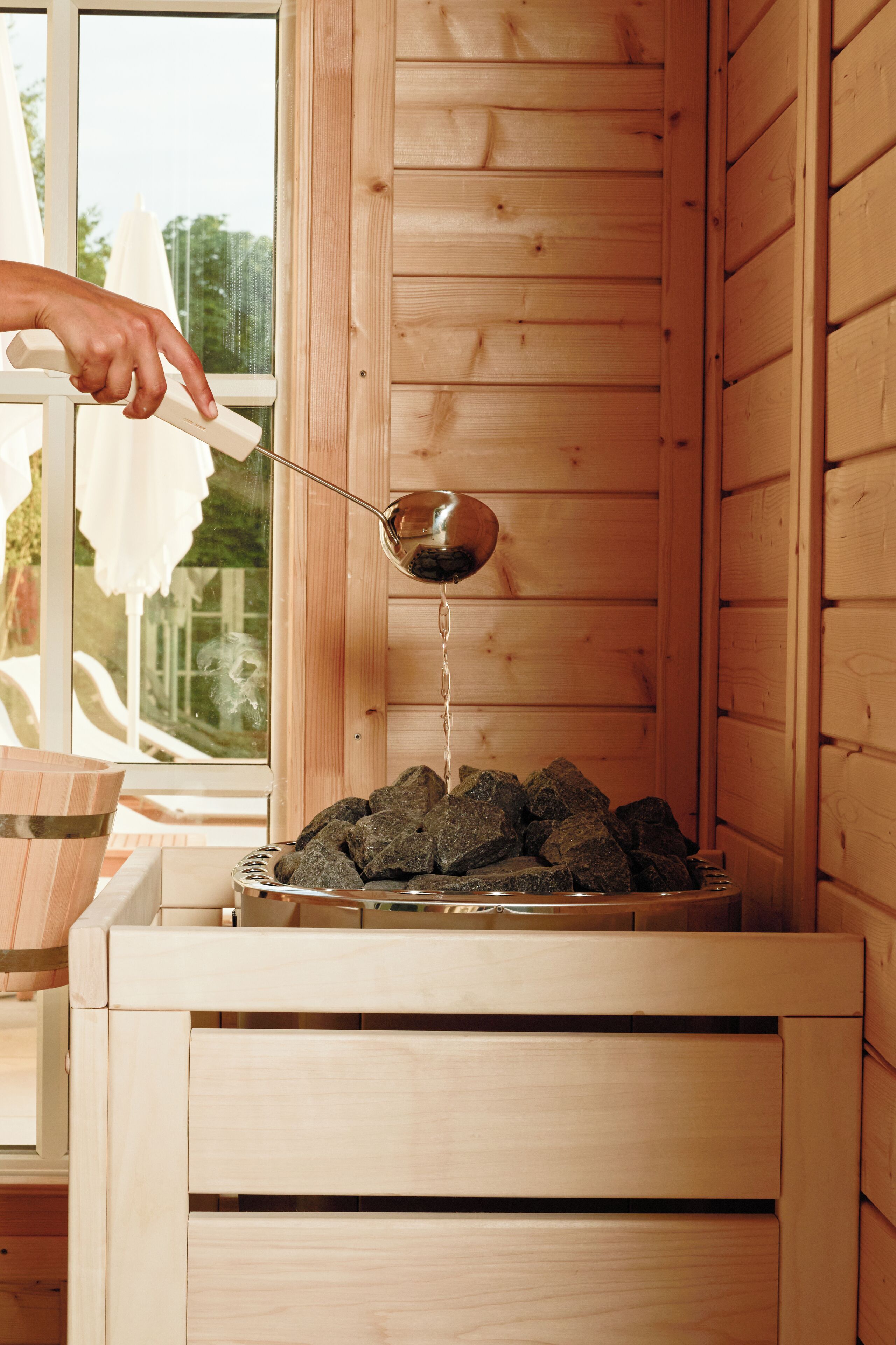 Un sauna avec des murs en bois et une fenêtre, une personne versant de l’eau sur des pierres, et un banc en bois.