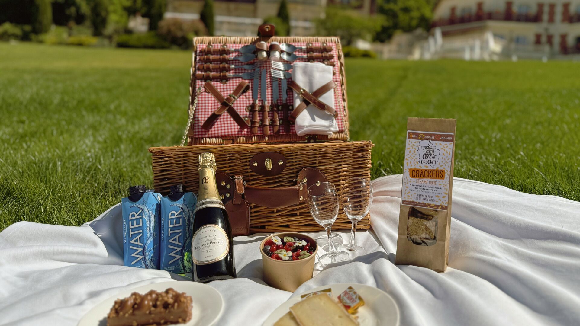 A picnic setup on a grassy field with a wicker basket containing a bottle of champagne, wine glasses, a bowl of fruit, and a bag of crackers.