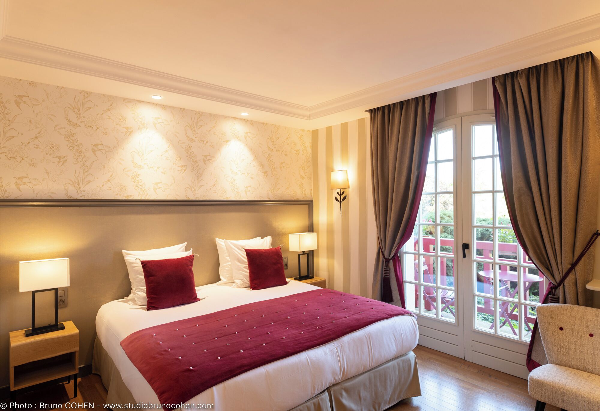 A hotel room with a large bed, two bedside lamps, a nightstand, a chair, and a view of the outside through a window with curtains.