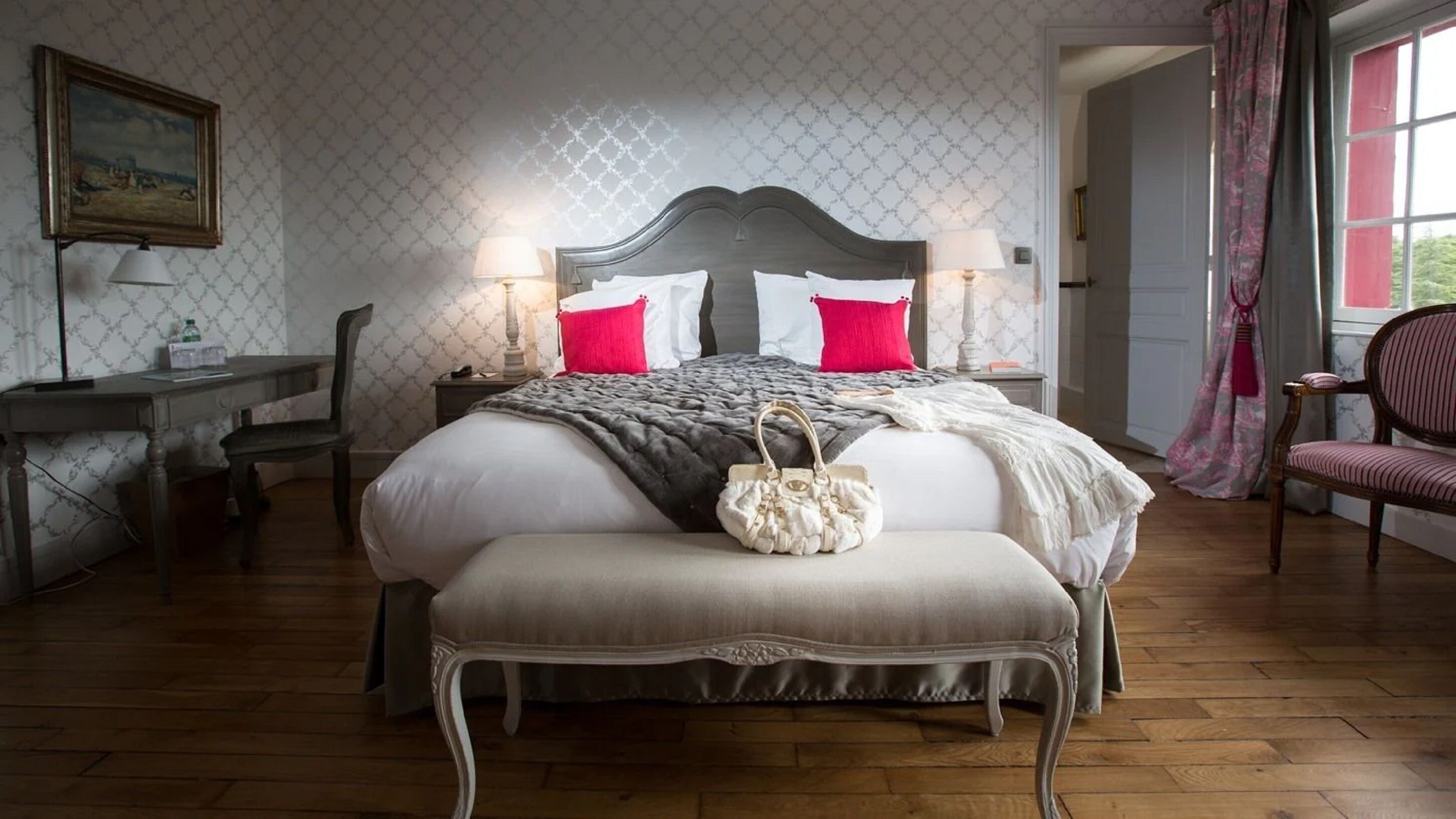 A bedroom with a large bed, two bedside tables with lamps, a desk, a chair, and a window with curtains.