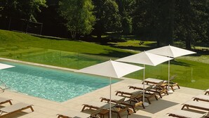 Une piscine entourée d’une pelouse et d’arbres, avec des chaises longues et des parasols à proximité.