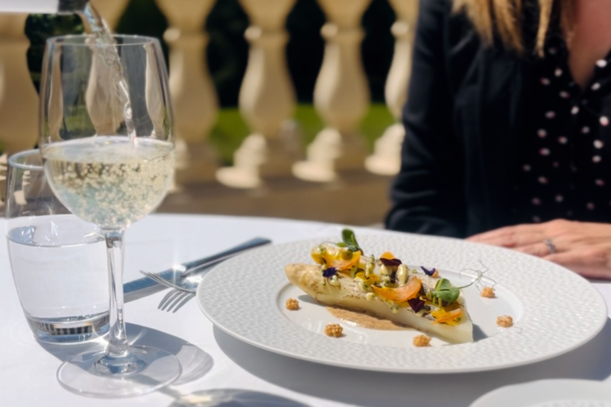 Una mesa de comedor con un plato blanco de comida, una copa de vino blanco y cubiertos.