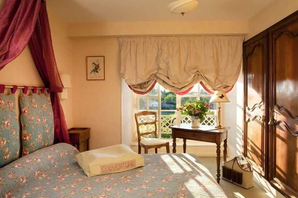 A bedroom with a floral bedspread, a wooden wardrobe, a window with curtains, and a small table with a lamp.