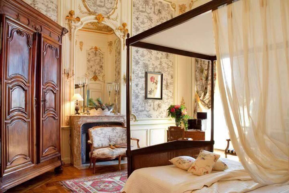 A bedroom with a four-poster bed, a wooden wardrobe, a fireplace, and floral wallpaper.