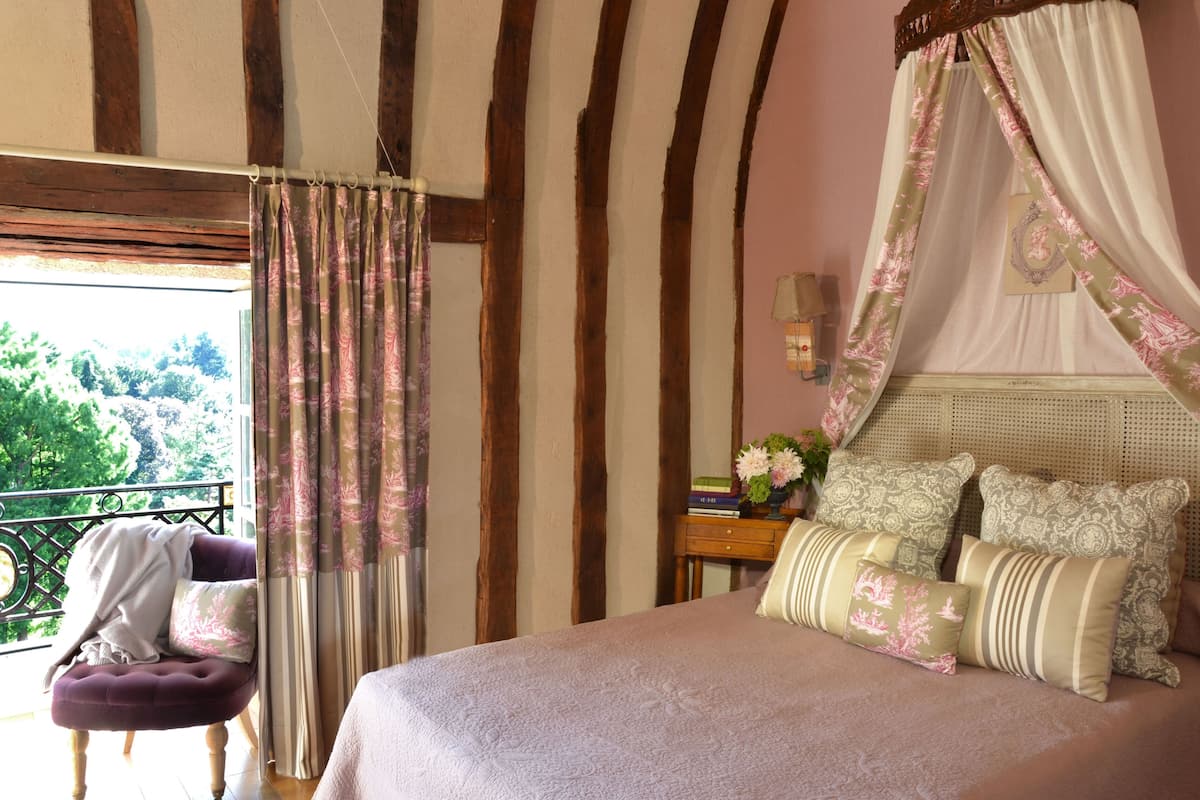 A bedroom with a four-poster bed, a nightstand with books and a vase of flowers, and a view of greenery through a window.