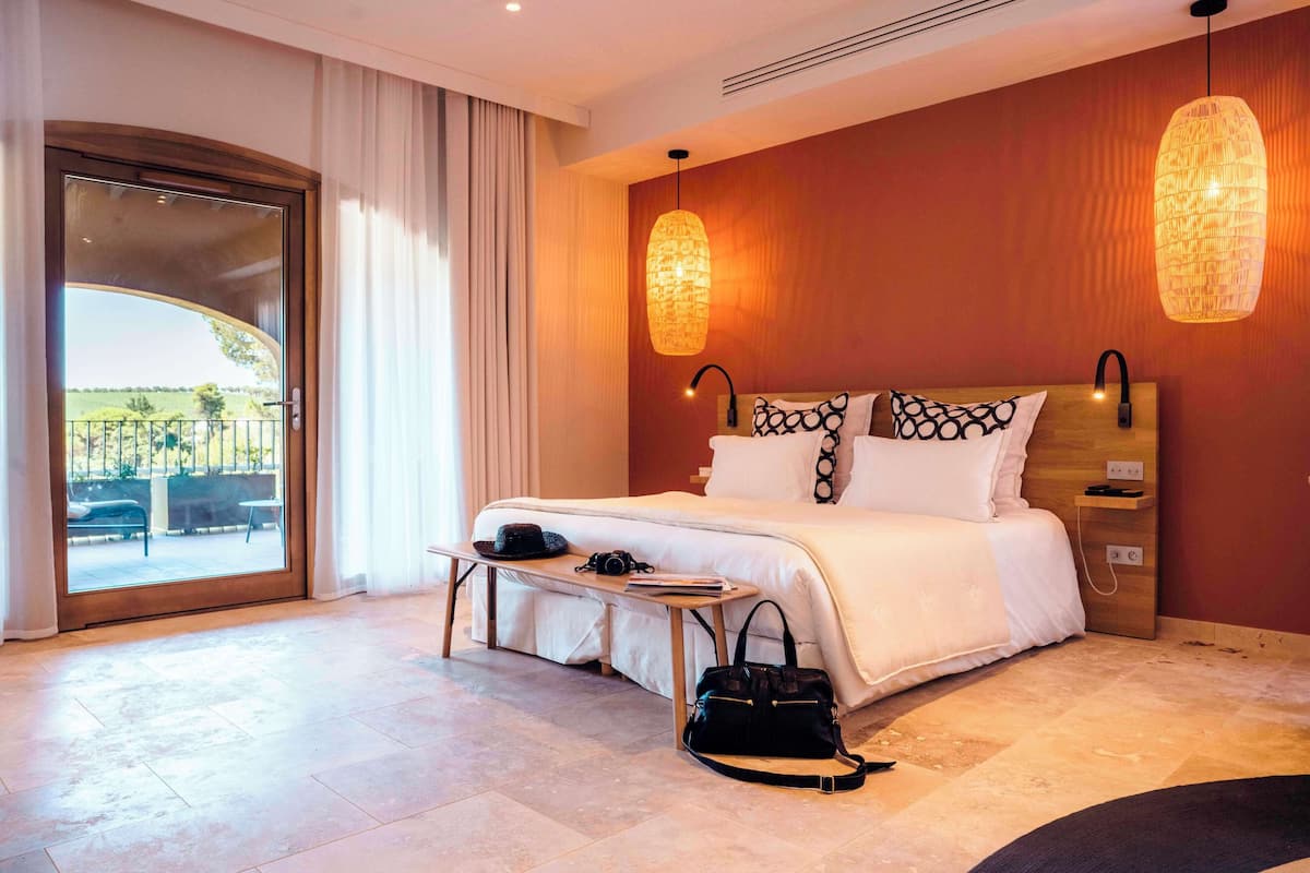 Ein Hotelzimmer mit einem großen Bett, einem Schreibtisch, einem Stuhl, einer schwarzen Handtasche und Blick durch eine Glastür.
