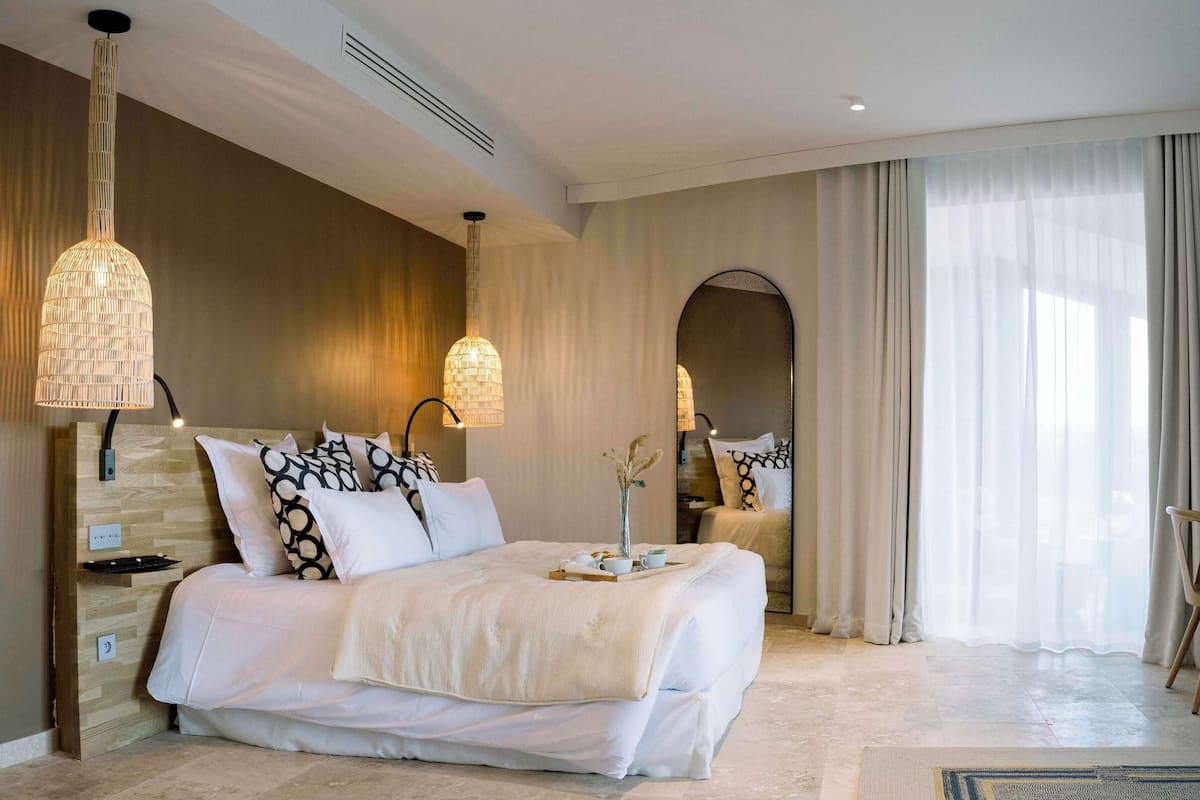 Ein modernes Schlafzimmer mit einem großen Bett, zwei Wandleuchten, einem Spiegel und Blick ins Freie durch transparente Vorhänge.