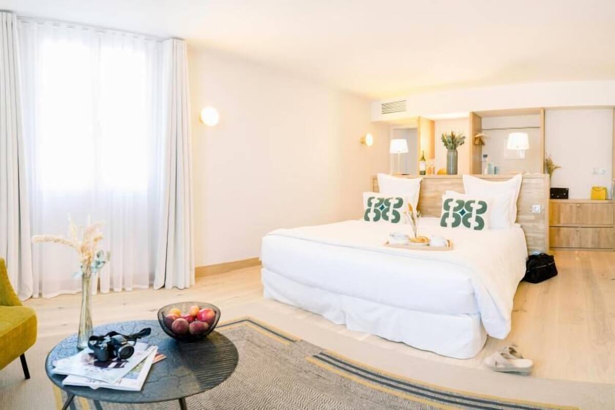 Ein Hotelzimmer mit einem Bett, einem kleinen runden Tisch mit einer Obstschale, einem Sessel und einem Nachttisch mit Lampe und einer Vase.