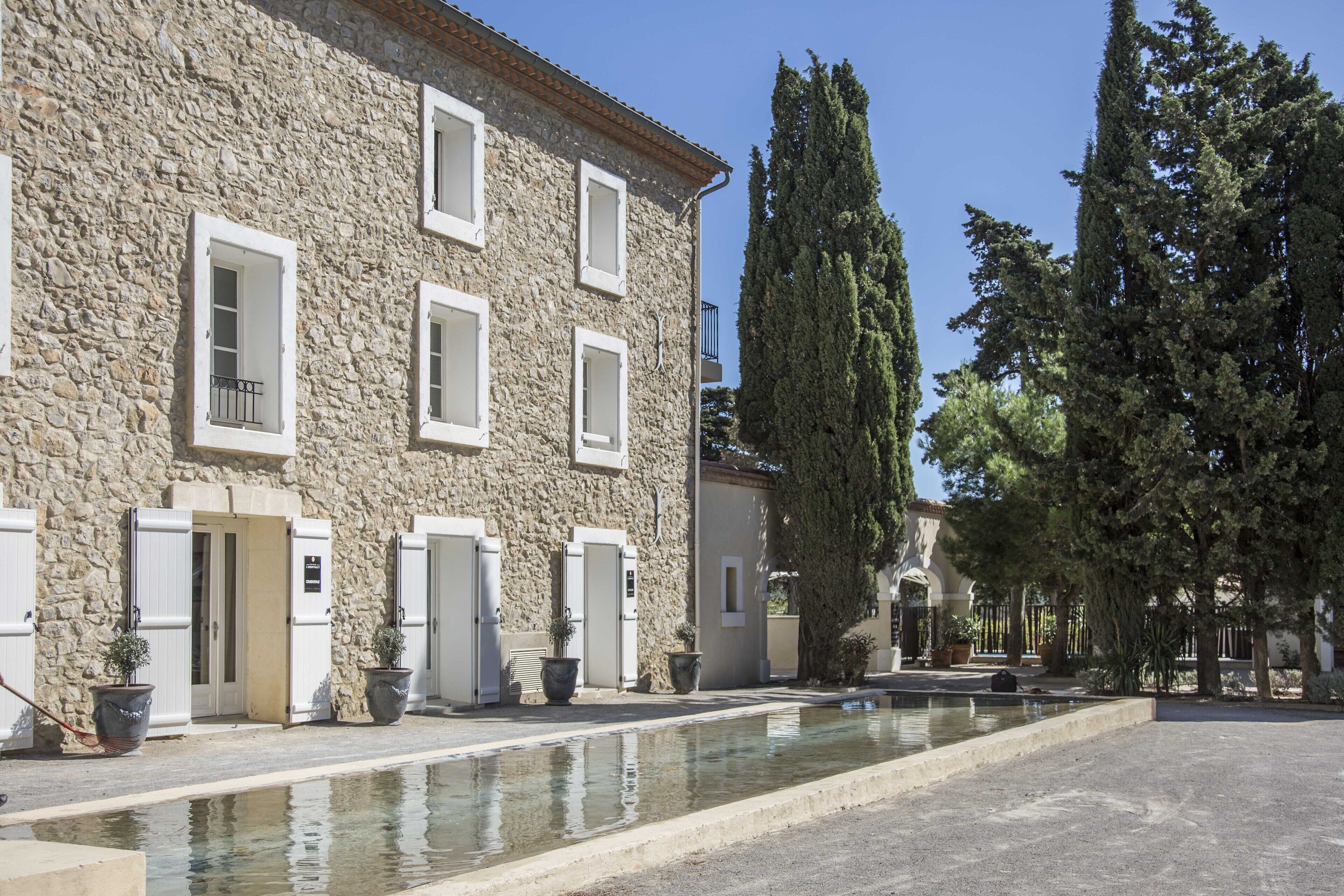 Un bâtiment en pierre doté de nombreuses fenêtres et portes, une cour avec un élément décoratif aquatique et de grands arbres.