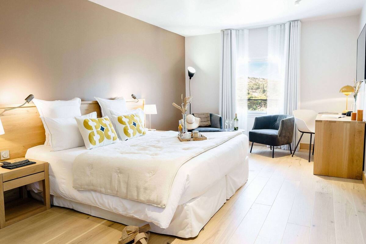 Ein Hotelzimmer mit einem großen Bett, Nachttischen, einem Schreibtisch, einem Sessel und Blick ins Freie.
