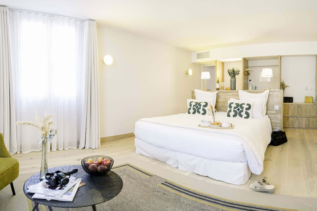 Ein Hotelzimmer mit einem großen Bett, einem runden Tisch mit Obstschale, einem Sessel und einem Spiegel.