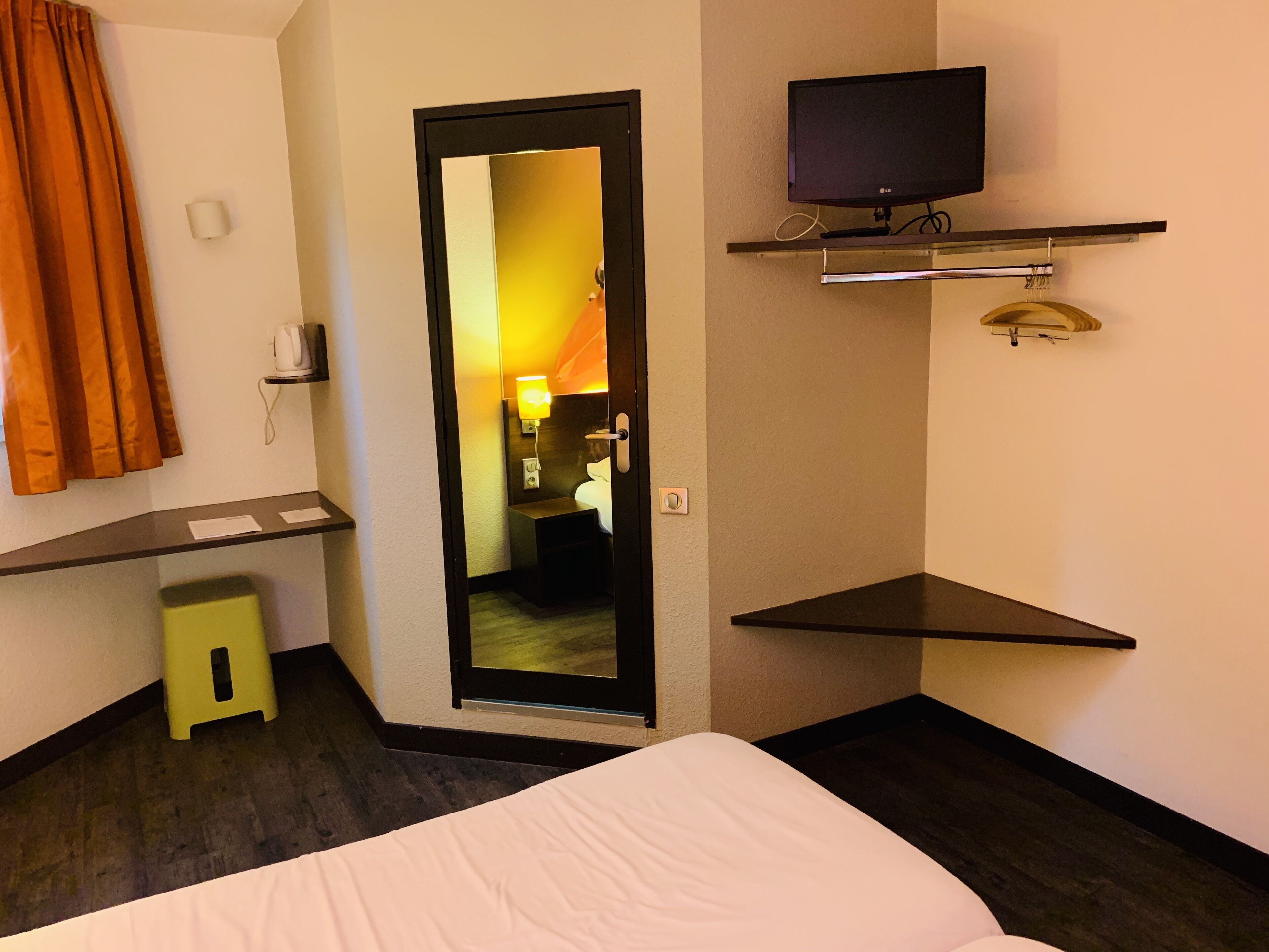 A hotel room with a bed, a small stool, a mirror, a TV on a shelf, and a door leading to another room.
