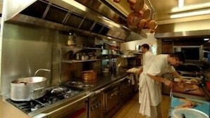 Una cocina comercial con electrodomésticos de acero inoxidable, un chef preparando comida y ollas de cobre colgando.