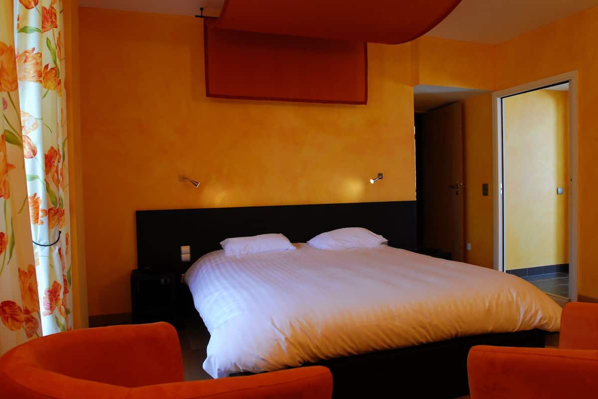 A hotel room with a bed, two orange chairs, a nightstand, and a door leading to another room.